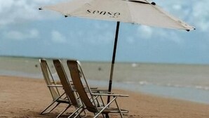 En strandstol och ett parasoll med ordet ”sense” på sig, placerade på en sandstrand med utsikt över havet och himlen.