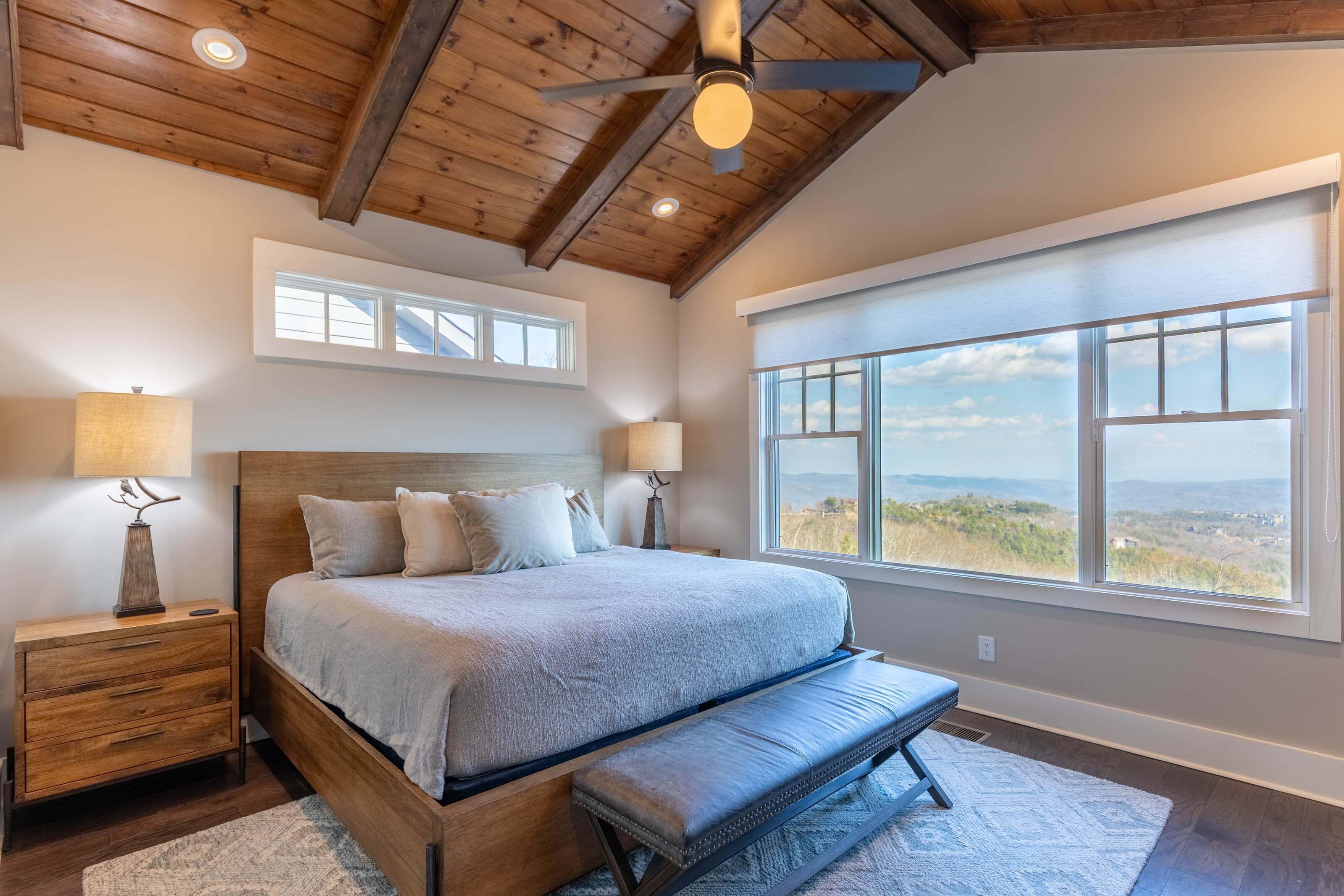 A bedroom with a large bed, wooden headboard, two bedside lamps, a ceiling fan, and a large window with a view.