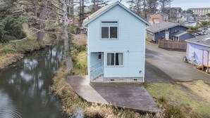 A light blue house with a small porch, a narrow waterway, and a forested area in the background. - Canal Cottage with Kayak Dock, Walk to Beach (Lincoln City)