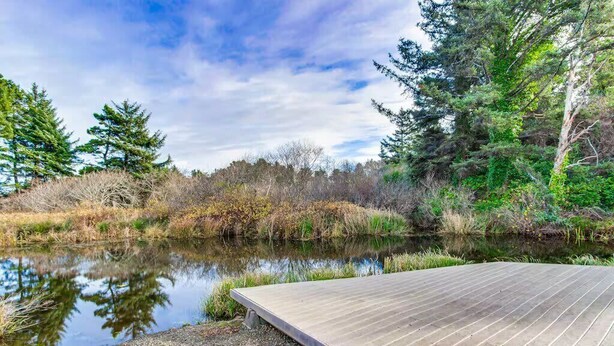 A wooden deck extending over a calm body of water, surrounded by dense trees and shrubs. - Canal Cottage with Kayak Dock, Walk to Beach (Lincoln City)