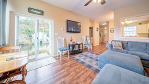 A living room with a wooden dining table, two chairs, a blue sofa, a ceiling fan, and a TV mounted on the wall. - Canal Cottage with Kayak Dock, Walk to Beach (Lincoln City)
