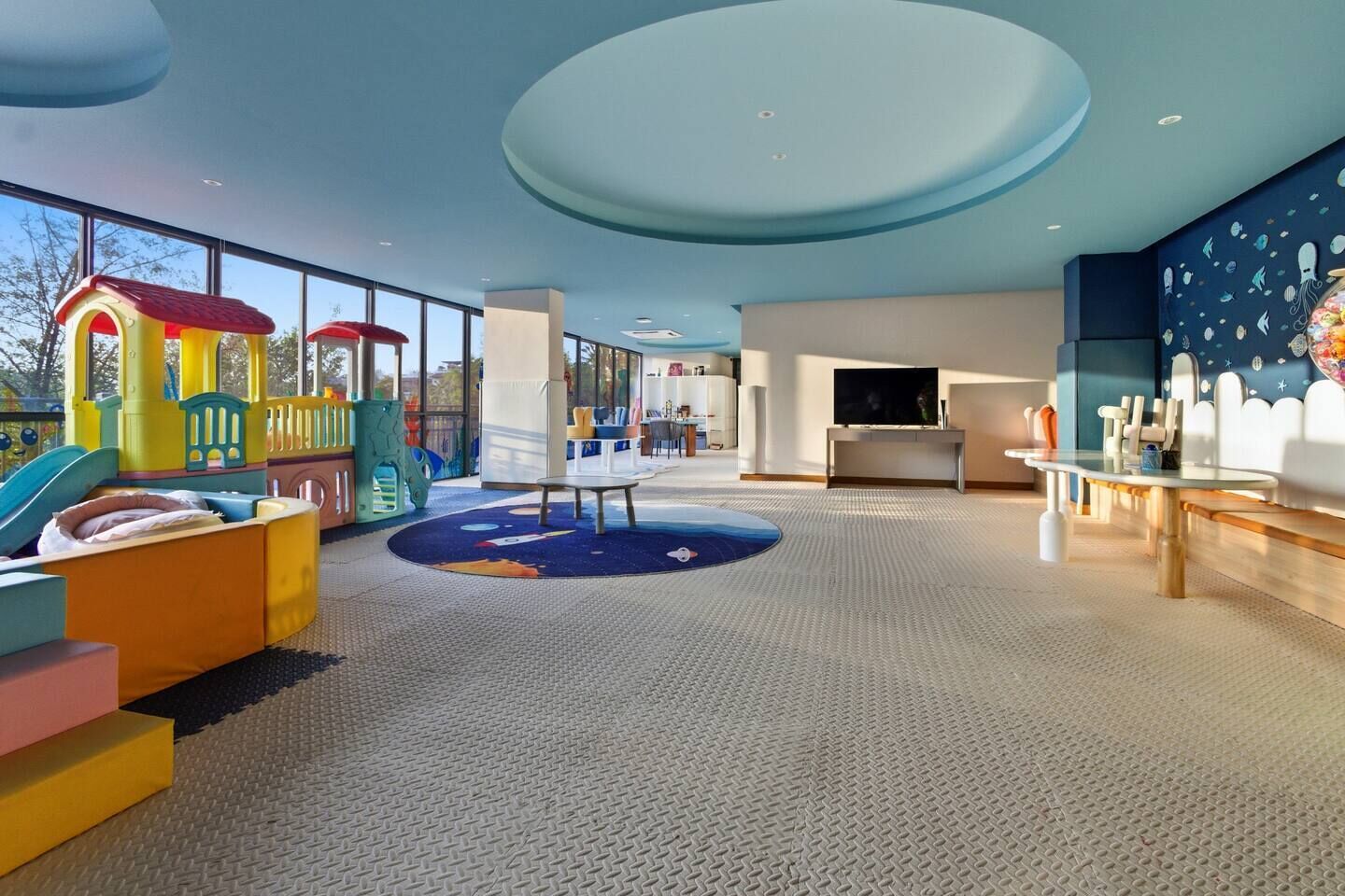A children's play area with a slide and climbing structures, a seating area with a table, and a large window providing a view of the outdoors.