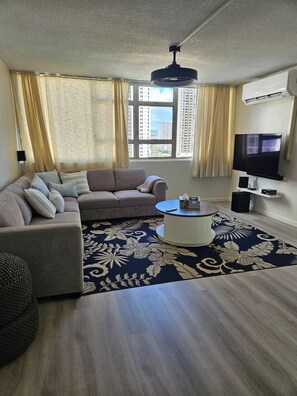 A living room with a sofa, a coffee table, a TV, and a large window with curtains. - Waikiki  beach is just across  street. (Honolulu)