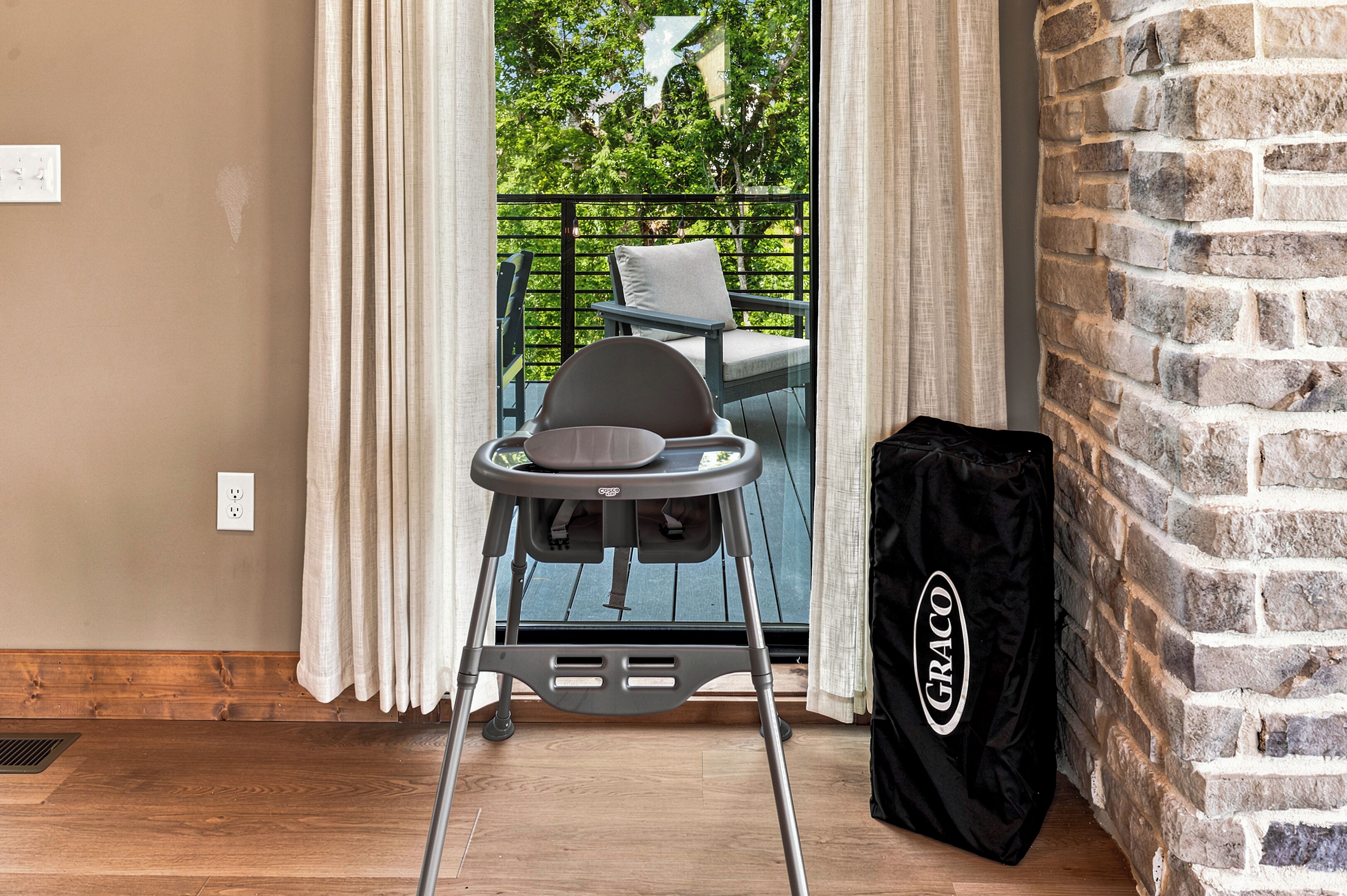 Une pièce avec un plancher en bois, un sac noir sur lequel on peut lire le mot « GRACO », une chaise avec un coussin et une vue sur la verdure à travers une porte-fenêtre donnant sur un balcon.