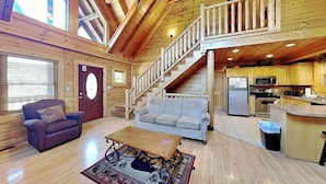 A spacious living room with a sofa, a coffee table, and a staircase leading to a balcony. - Budget friendly, Smoky Mountain Cabin Rental | Sleeps 10 | Pool Table (Pigeon Forge)