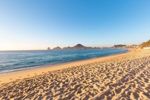 Une vaste plage de sable fin, avec des vagues douces, un ciel dégagé et des formations rocheuses lointaines.