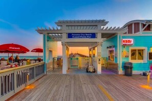 A pier with a Coca-Cola branded umbrella, a sign for Panama City Beach, and a building with a neon 'OPEN' sign. - Coastal Luxe 1024-Gulf Views-Sleeps 8 (Panama City)
