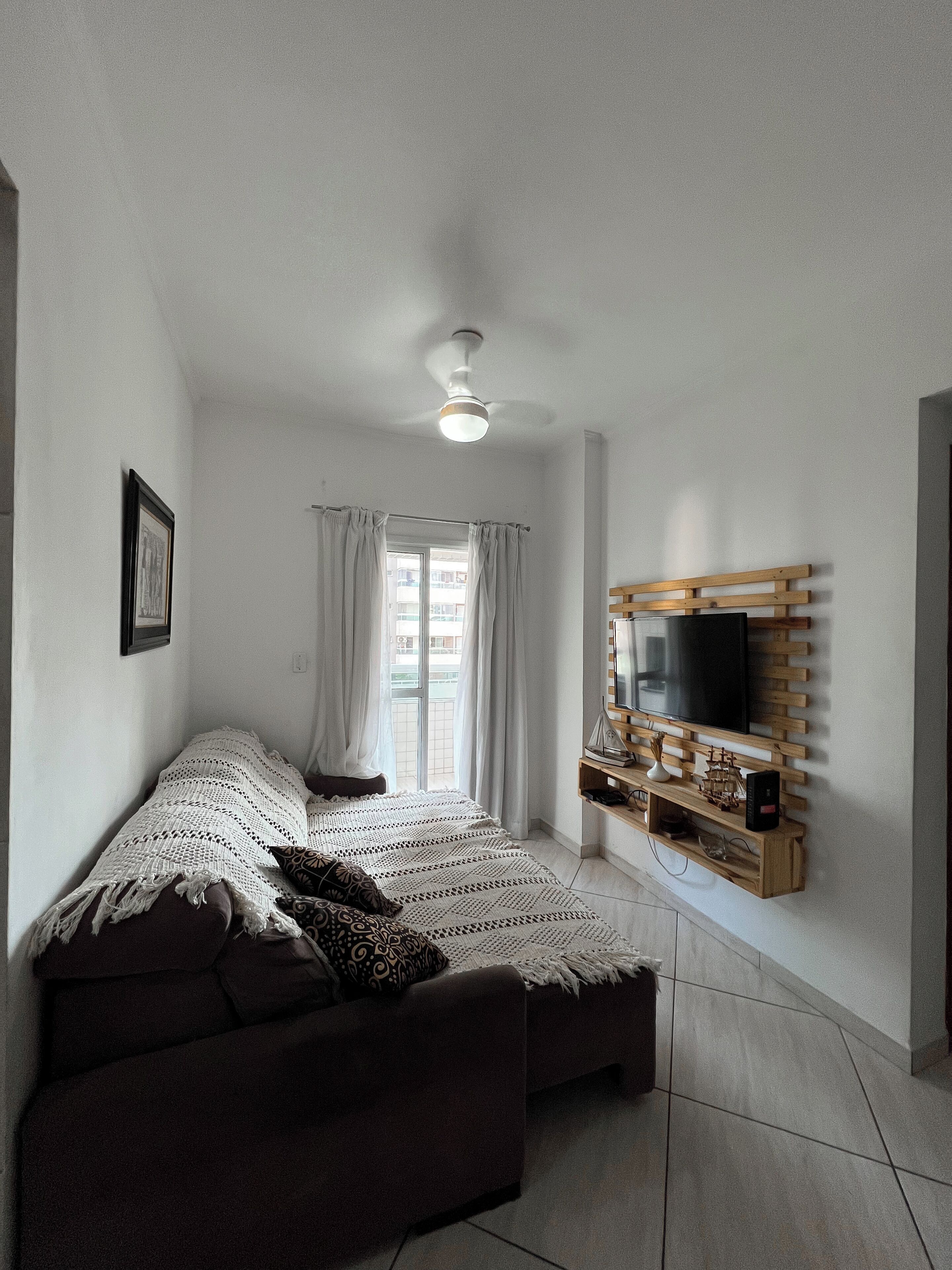 A bedroom with a bed, a ceiling fan, a TV mounted on a wooden shelf, and a window with curtains.
