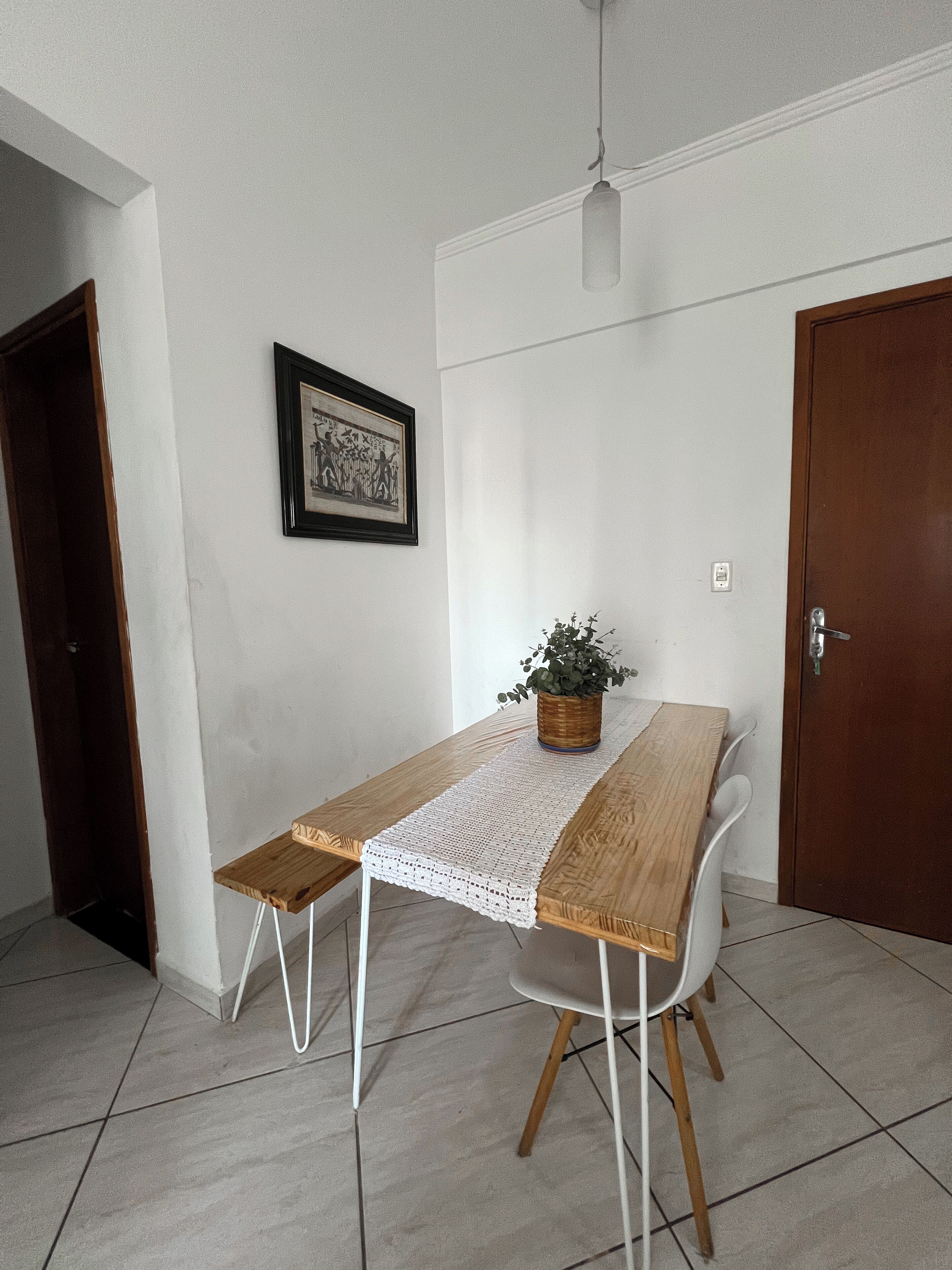 A dining table with a woven table runner, a potted plant, and a framed picture on the wall.