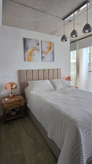 Un dormitorio moderno con una cama grande, dos lámparas colgantes fijadas a la pared, una mesita de noche con una lámpara y arte abstracto en la pared.