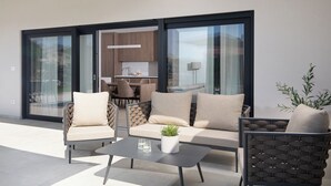 Een terras met een bank, stoelen en een tafel, een keuken zichtbaar door grote glazen deuren, en een plant in een pot op de tafel.
