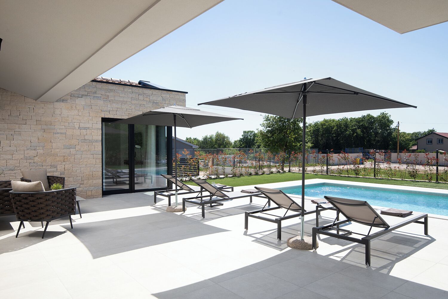 Un área de piscina moderna al aire libre con sillones de descanso y sombrillas, una pared de piedra y un vidrio que da a un porche cubierto.