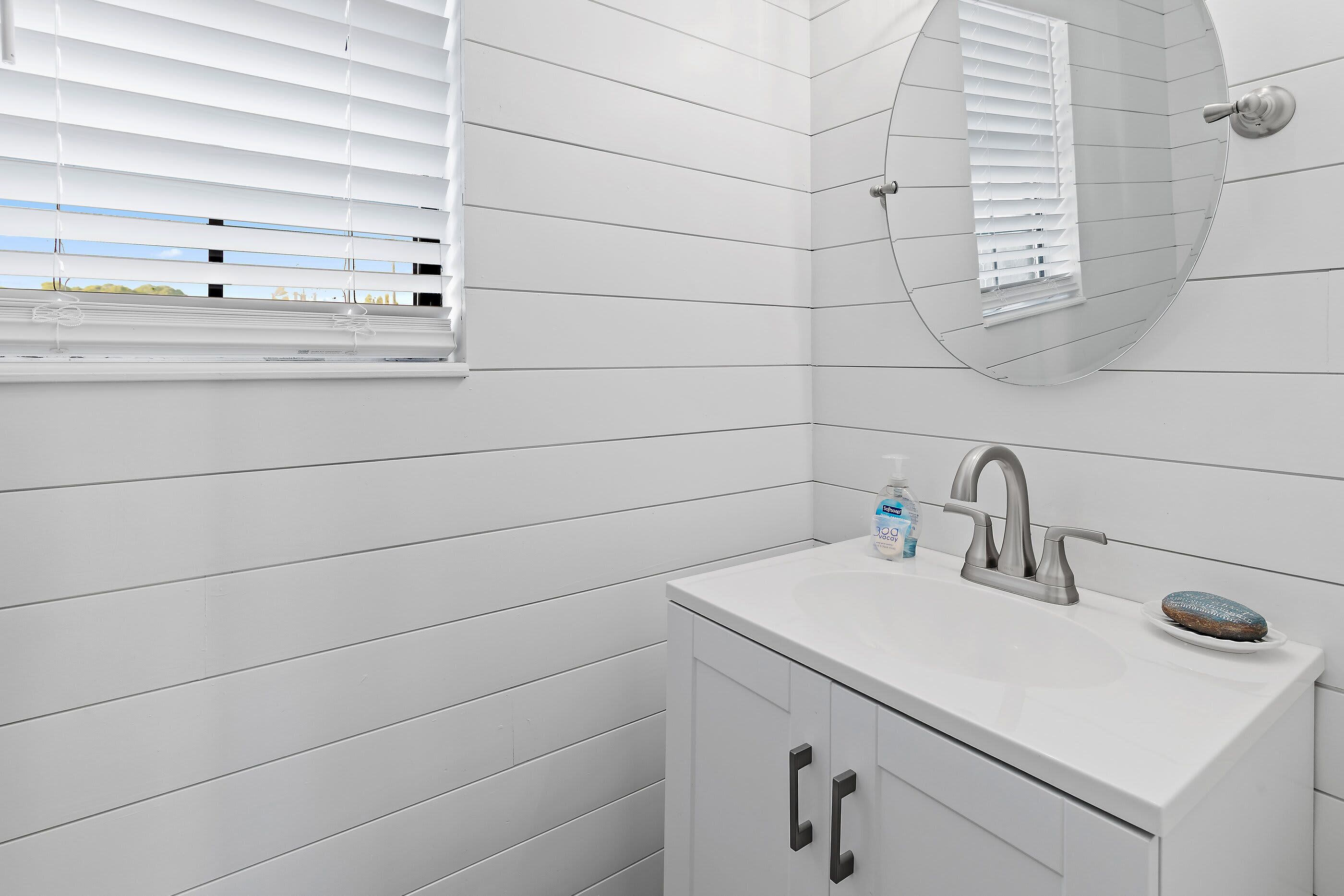 A bathroom with white tiled walls, a white sink, a mirror, and a window with blinds.
