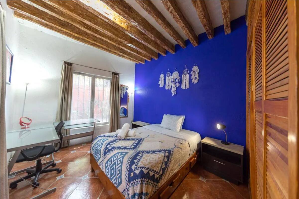 A bedroom with a bed, a desk, a chair, a window, and a blue wall.