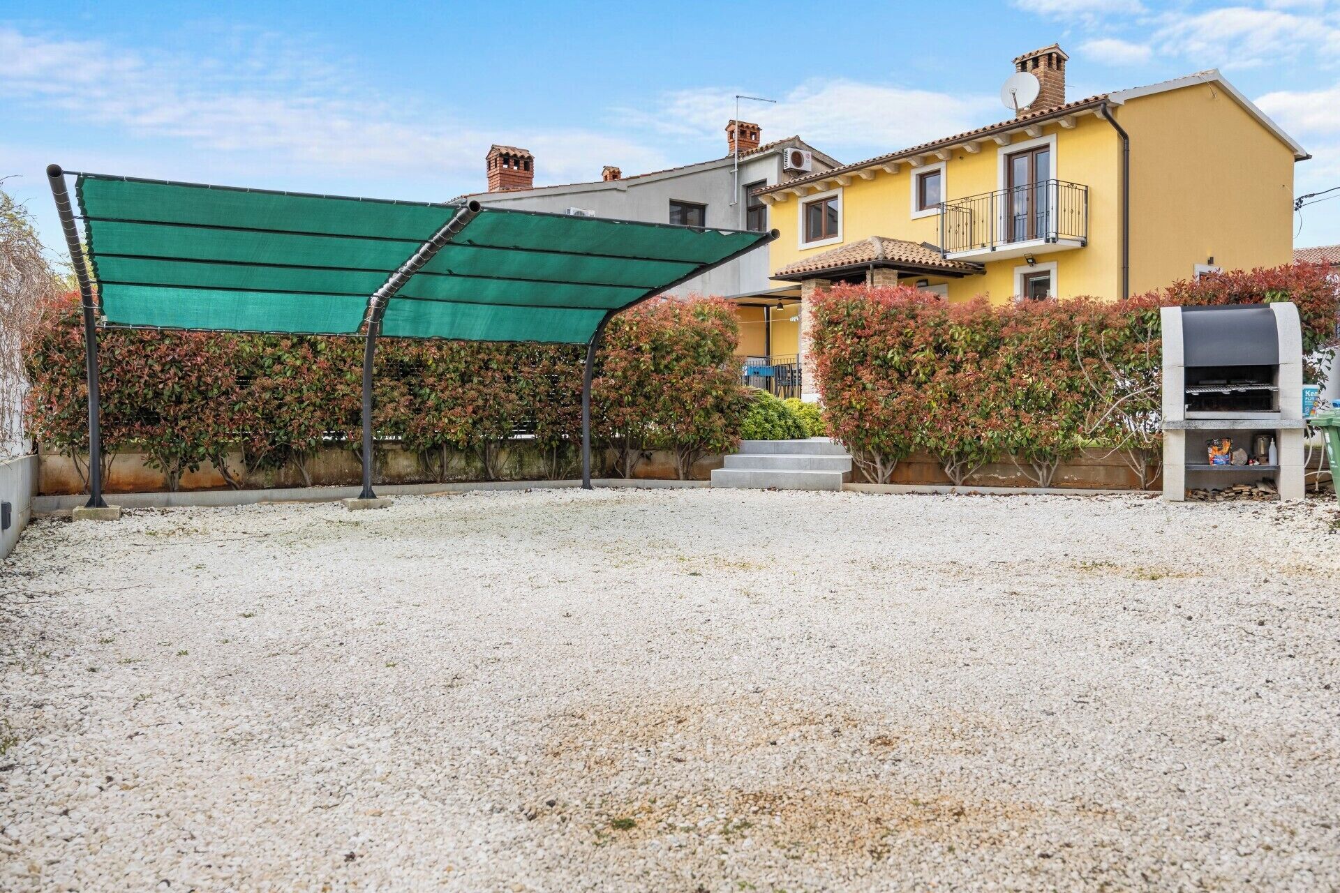 Zona residencial con camino de grava, follaje verde y un edificio amarillo con balcón.