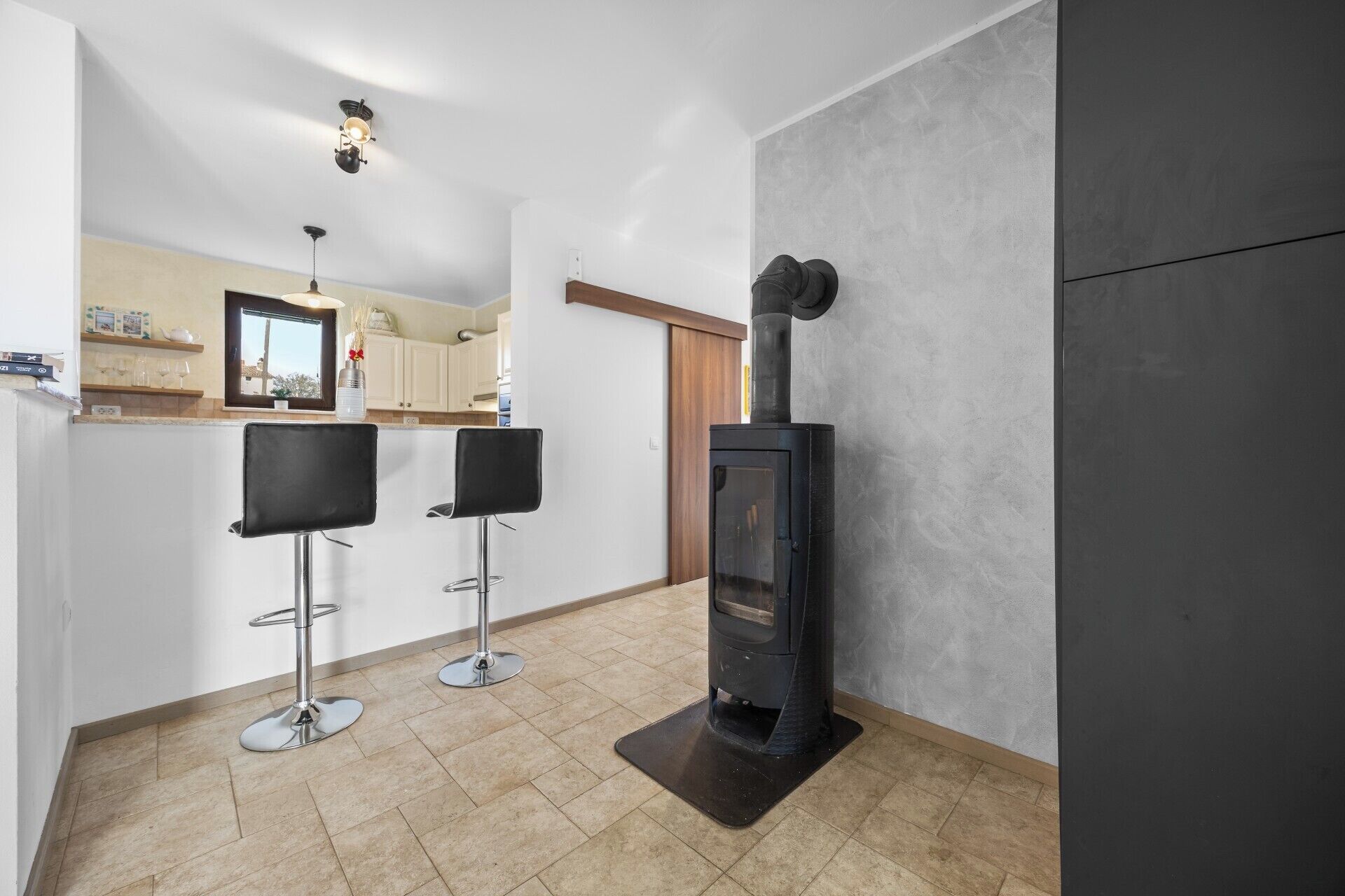 Eine moderne Küche mit einem schwarzen Kaminofen, zwei Barhocker und einer Kücheninsel mit Fenster.