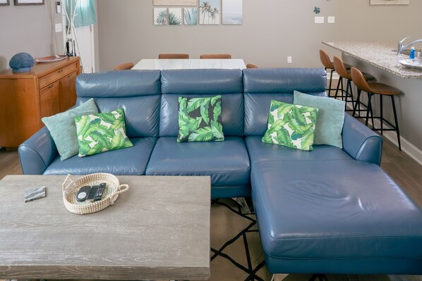 Ein blaues Ledersofa mit grün gemusterten Kissen, einem Holztisch und einem gewebten Korb mit Fernbedienung.