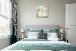 Ein ordentlich bezogenes Bett mit einer grün-weißen Streifenauflage, zwei Nachttischlampen, einem Kissen mit dem Schriftzug 'relax' und einem Wandbild im tropischen Stil.