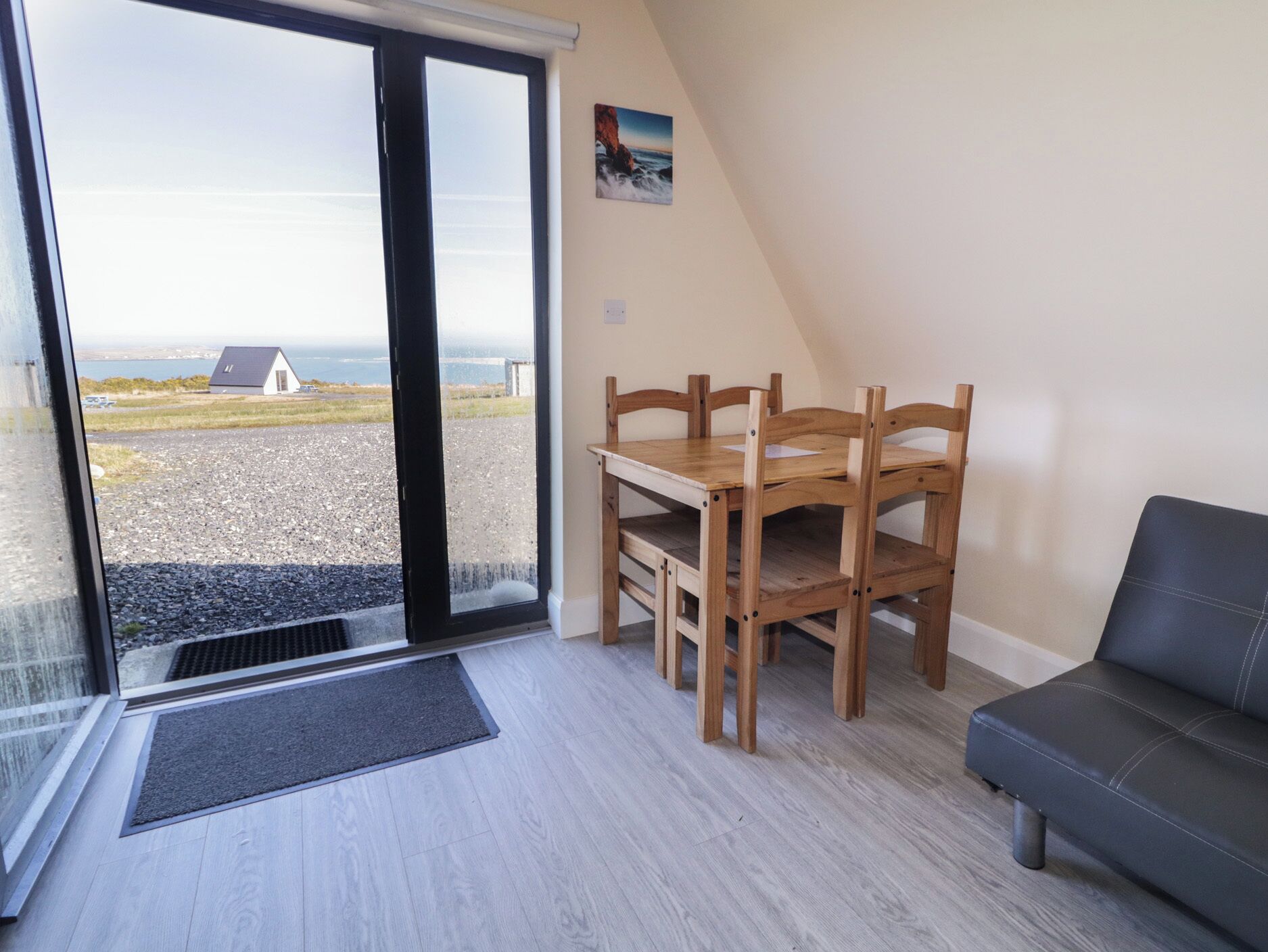 Een kamer met een houten eettafel en stoelen, een zwarte leren bank, uitzicht door een glazen deur naar buiten, en een klein schilderij aan de muur.