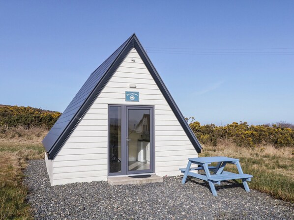 A small, white, triangular building with a dark roof and a door. There's a sign above the door and a blue bench in front.