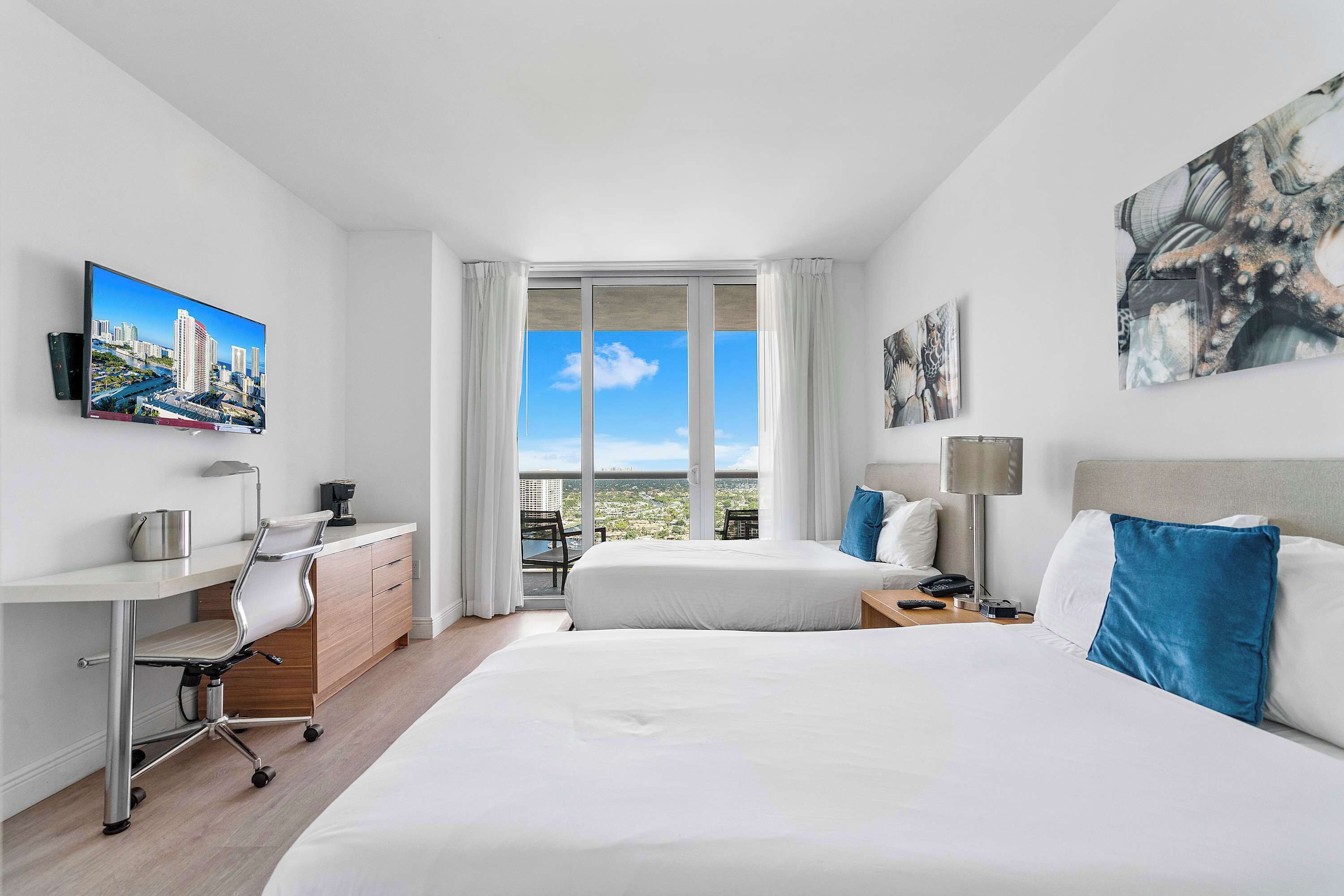 A hotel room with a bed, desk, chair, and a large window with a city view.