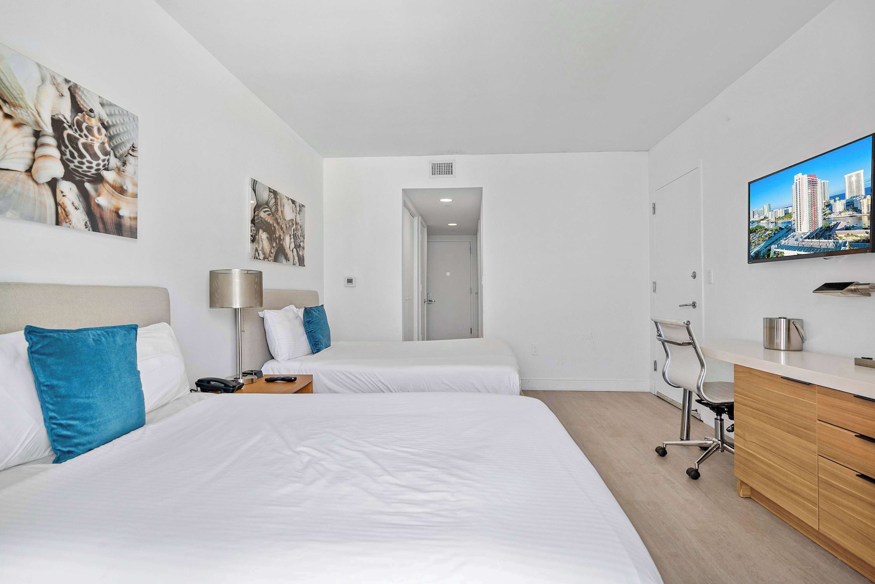 Une chambre d’hôtel avec deux lits, un bureau, une chaise, une télévision et des œuvres d’art accrochées aux murs.