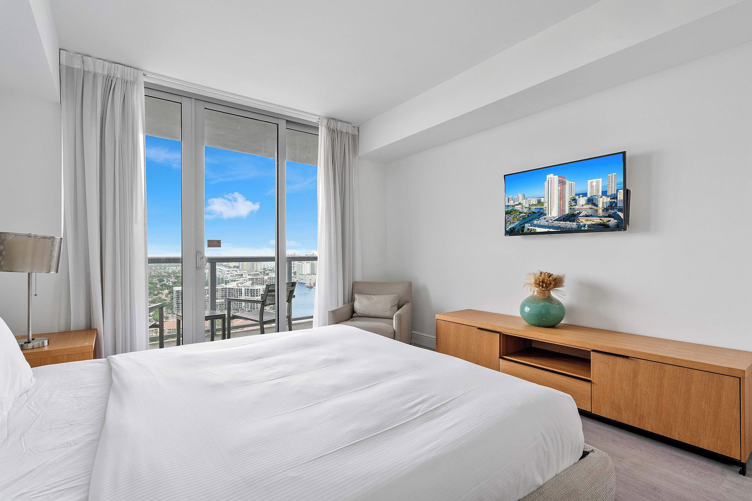 A hotel room with a large bed, a TV mounted on the wall, a wooden desk, a chair, and a view of the city through the window.