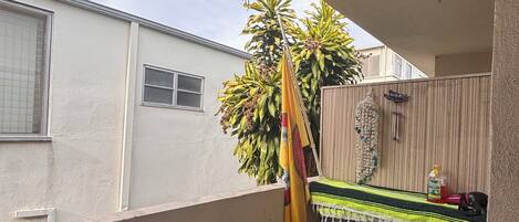 A small balcony with a green artificial turf floor, a wooden bench, a hanging wind chime, and a potted plant.