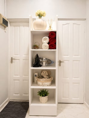A white interior with a shelf unit containing various items including a vase, rolled towels, a basket, and a potted plant.