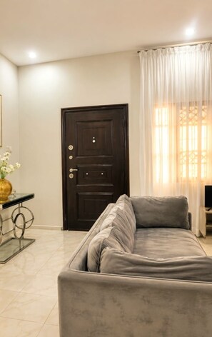 Una habitación con una puerta de madera oscura, un sofá gris, una mesita con un jarrón y una ventana con cortinas traslúcidas.