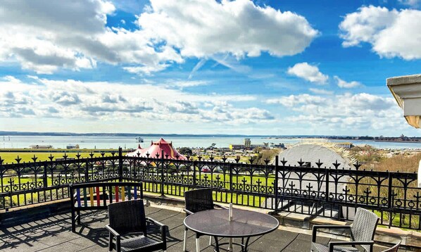Un balcon avec des sièges d’extérieur, une vue sur la mer et une structure rouge visible au loin.