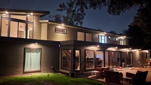 Una casa moderna de dos pisos amb grans finestres, un patio amb mobles d'exterior i una façana ben il·luminada.