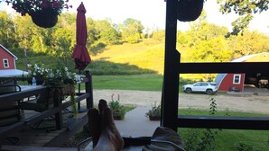 A wooden deck with a view of a green field, a red barn, and a white car parked outside.