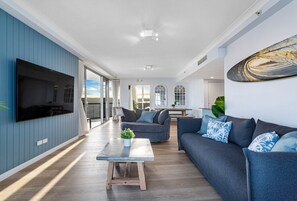 Ein modernes Wohnzimmer mit großem Flachbildfernseher, einer Couch, einem Couchtisch und einem großen Fenster.