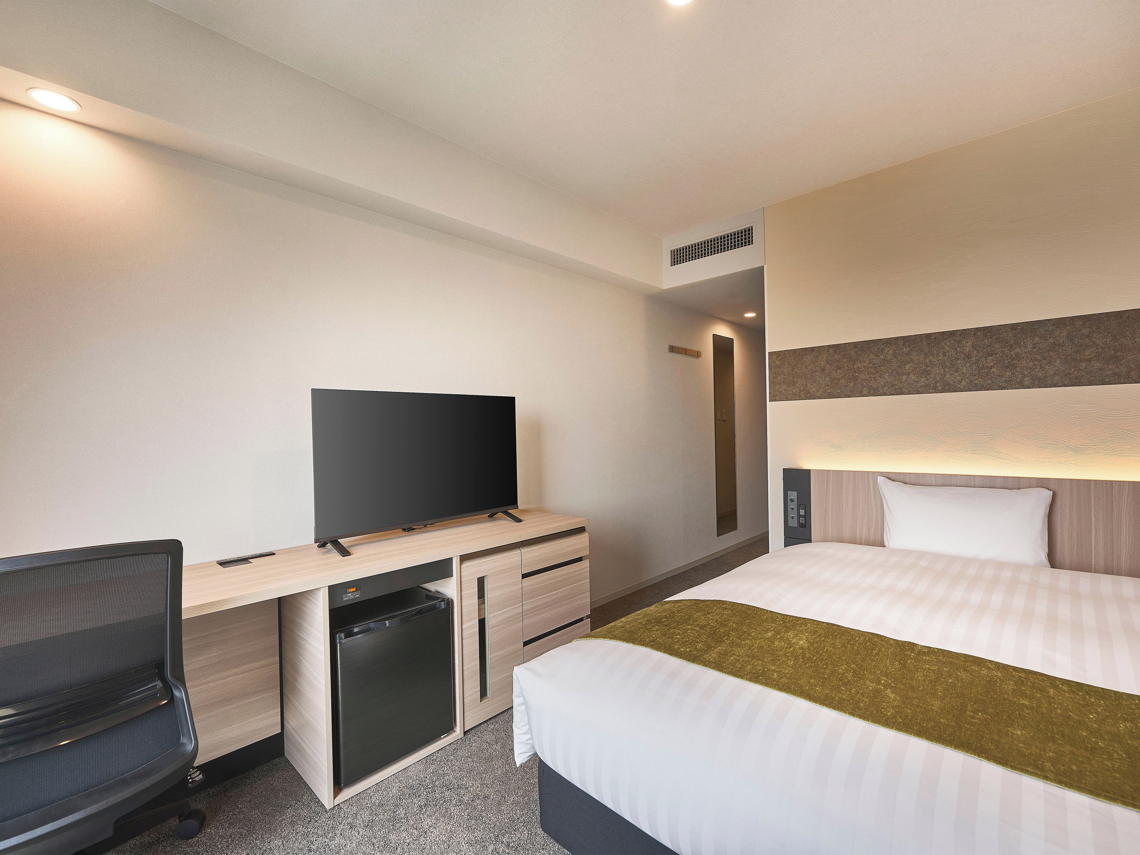A hotel room with a bed, a desk with a television, a chair, a small refrigerator, and a bedside table.