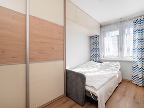 Une chambre à coucher avec un lit, une chaise grise, une fenêtre avec des rideaux et un plancher en bois.