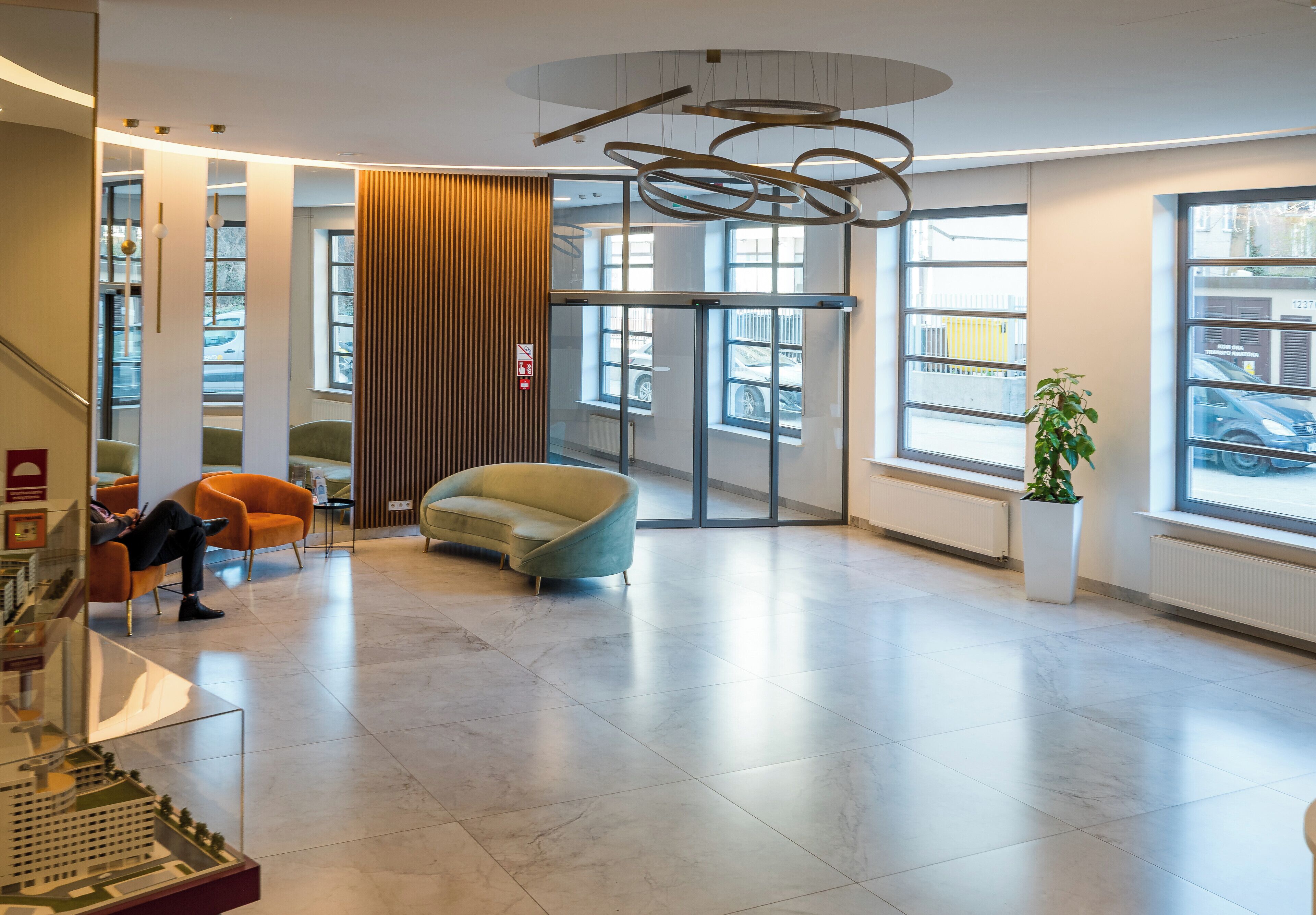 A modern lobby with a glass door entrance, a seating area with chairs, a potted plant, and a model display.
