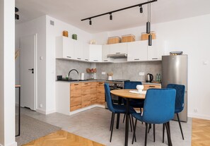 A modern kitchen with white cabinets, a stainless steel refrigerator, and blue chairs around a wooden table. - Krasiński Garden Skyline Apartment (Warszawa)