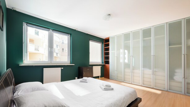 Een slaapkamer met een bed, twee ramen, een kledingkast en een radiator.