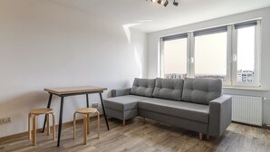 Ein modernes Wohnzimmer mit einem grauen Ecksofa, einem Holztisch mit zwei Hocker und einem großen Fenster mit Blick auf die Stadt.