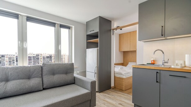 Ein kompakter Wohnraum mit grauem Sofa, einem Bett, einer Küchenzeile und Blick auf eine Stadtlandschaft durch große Fenster.