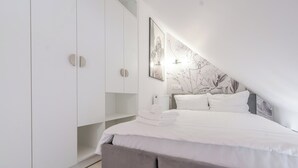 Un dormitorio con cama, cabecera gris, paredes blancas y un ventanal grande.