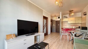 Ett modernt vardagsrum med en platt-TV, ett litet bord med fjärrkontroller, en matplats med stolar och ett kök med skåp och en kaffemaskin.