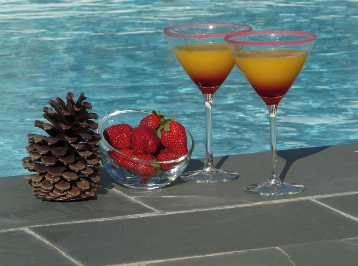 Dos vasos de martini con bebidas en capas y un bol de fresas junto a una piscina.