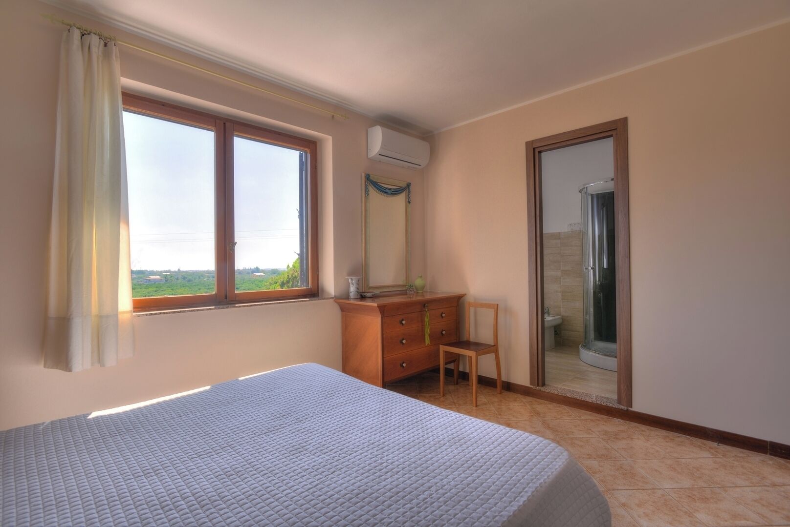 Une chambre à coucher comprenant un lit, une commode en bois, une chaise, une fenêtre avec des rideaux et une salle de bain avec une porte en verre.