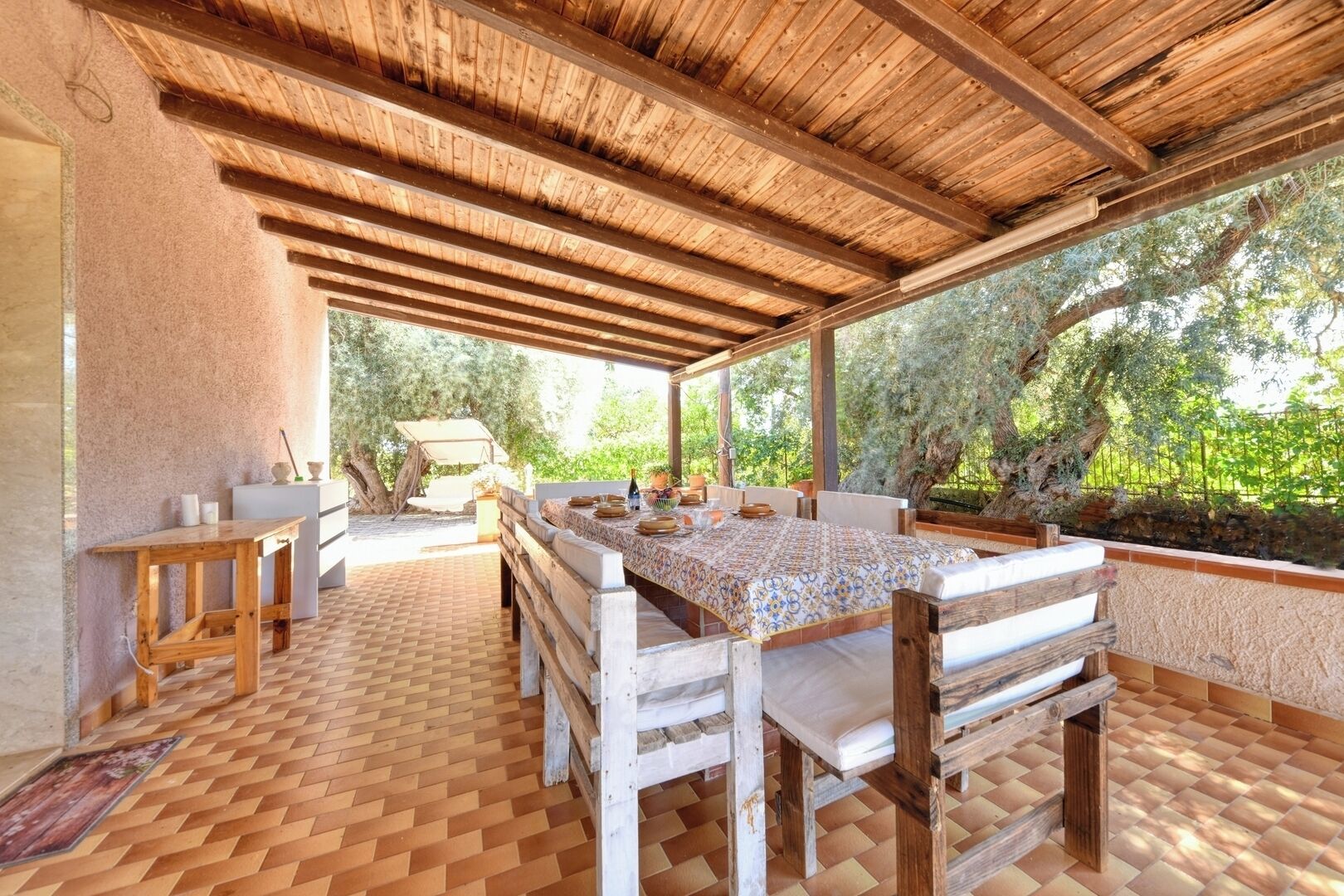 An outdoor covered patio with a dining table, benches, and a view of trees.