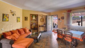 Een woonkamer met een bank, salontafel en een eettafel met stoelen. Een keuken is zichtbaar door een open deur.