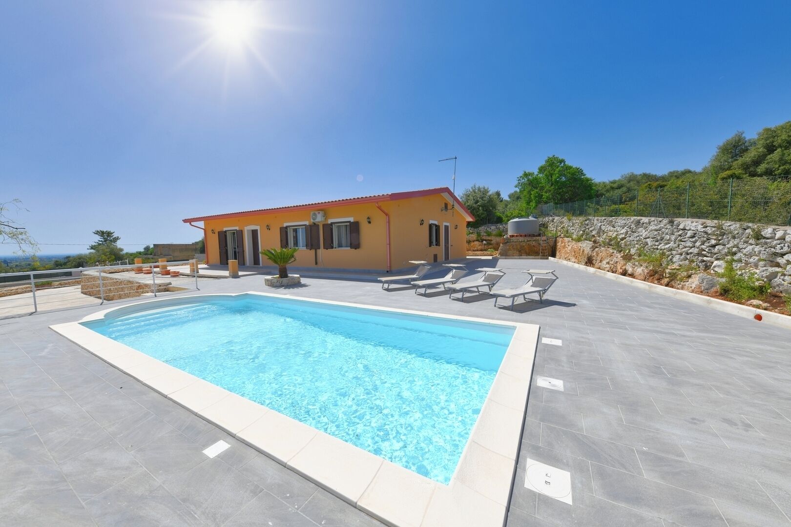 A swimming pool with lounge chairs, a small building with a red roof, and a stone wall in the background.