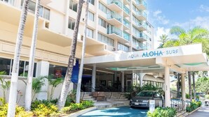 L’entrée d’un hôtel, avec un espace abrité, des palmiers et une enseigne indiquant « AQUA ALOHA SURF ».