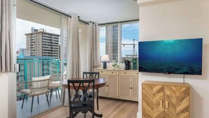 Une pièce comprenant une table à manger, des chaises, un téléviseur, un meuble et une vue sur les bâtiments, visible à travers une porte vitrée.